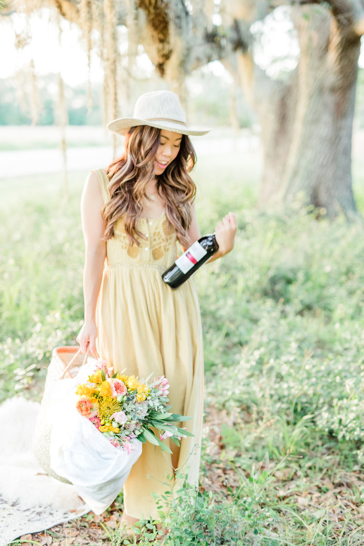 A woman examines a bottle of red wine while carrying a basket of fresh flowers outdoors