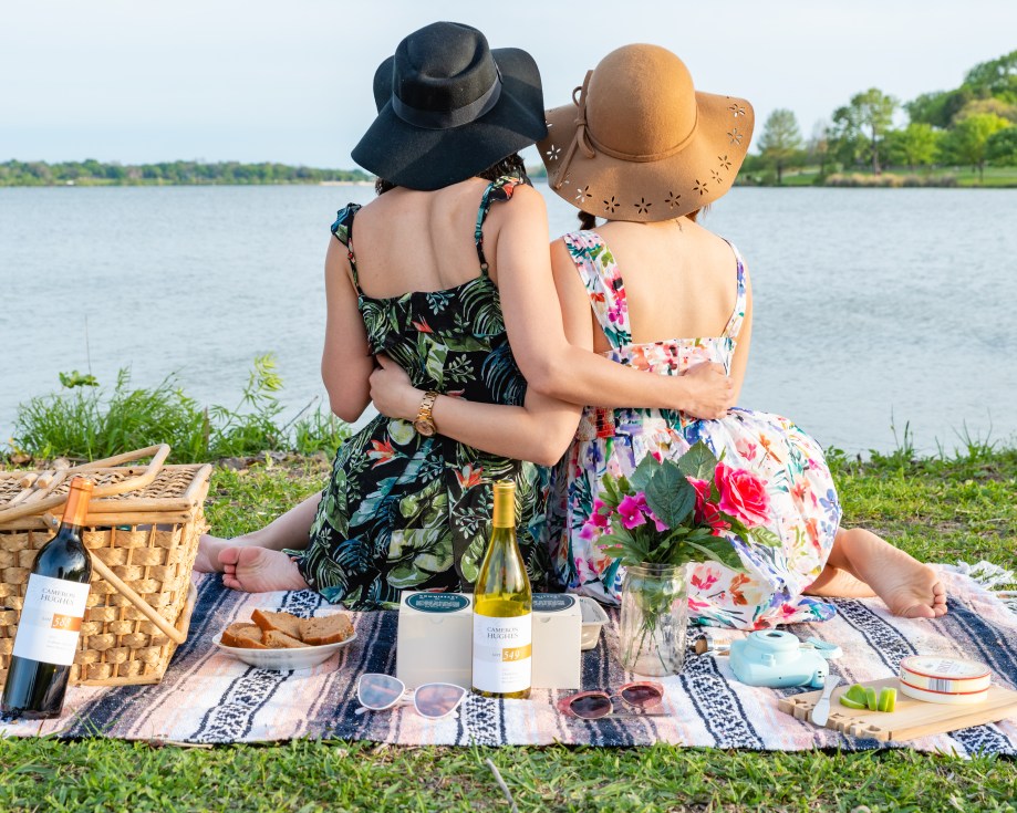 Two women embrace while enjoying a wine-paired picnic outdoors near a lake