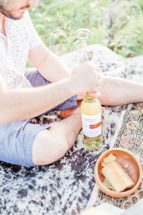 Man sits on a picnic blanket outdoors with a sandwich, opening a bottle of white wine