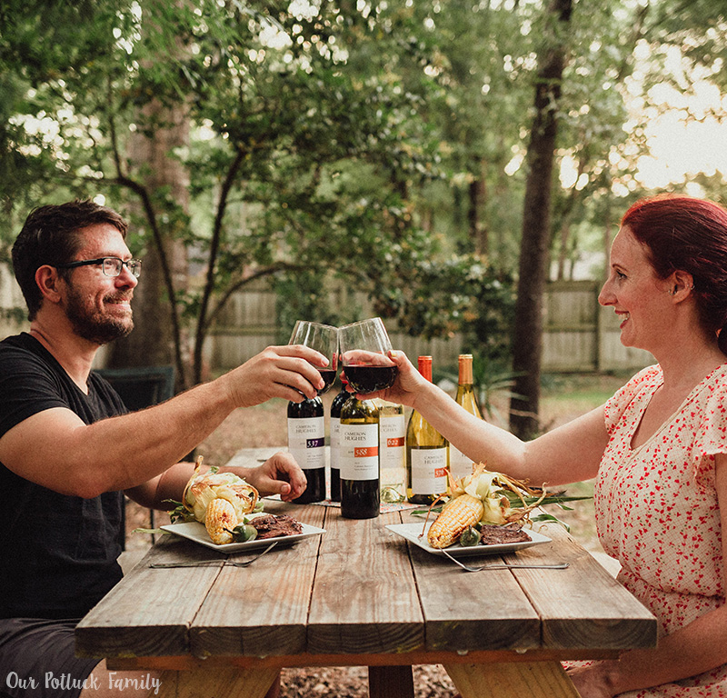 Man and woman cheersing with red wine outside while eating steak and corn
