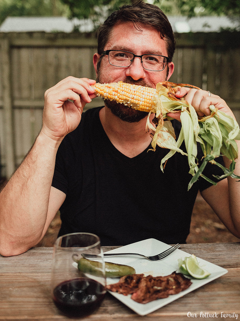 Man taking a bite out of corn on the cob with steak and wine nearby