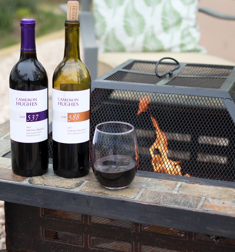 Two bottles of red wine with a glass poured and a fired up grill in the background