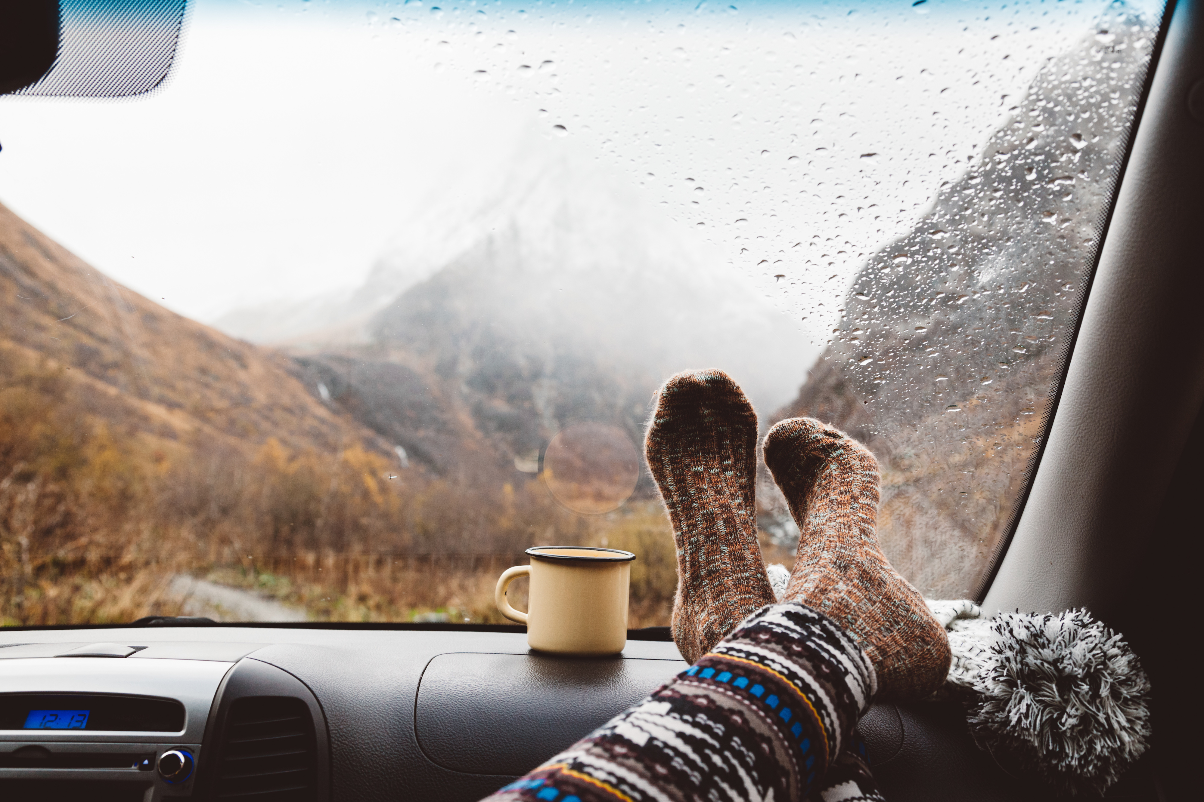 Woman legs in warm socks on car dashboard. Drinking warm tee on the way. Fall trip. Rain drops on windshield. Freedom travel