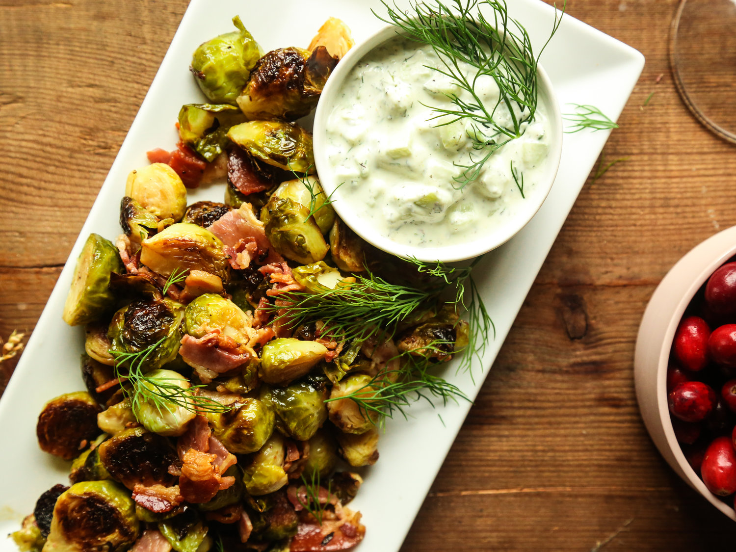 Roasted Brussels Sprouts with Pancetta and Tzatziki on a wood table next to a bowl of fresh cranberries
