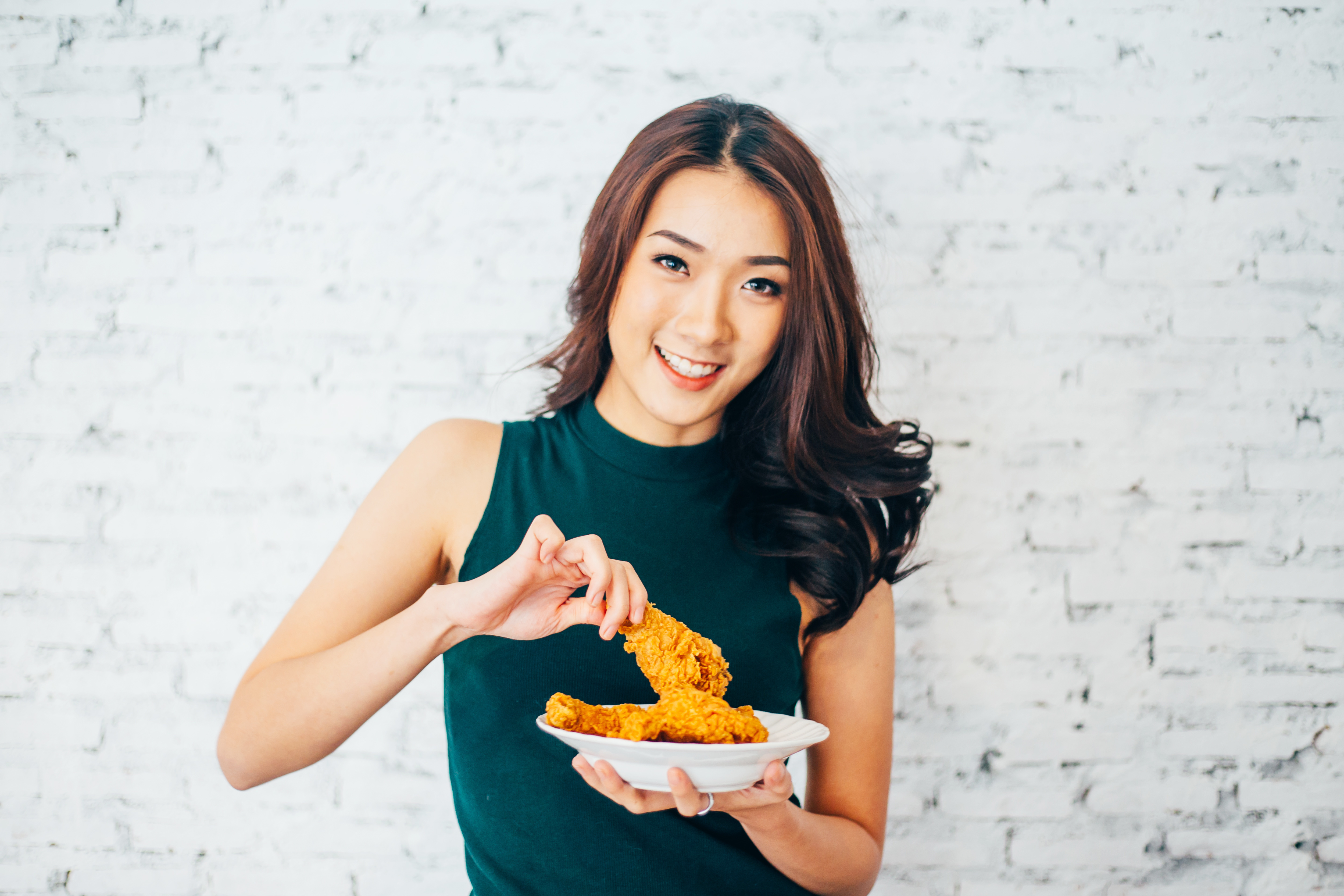 Attractive Asian woman eating fried chicken drumstick over white brick background