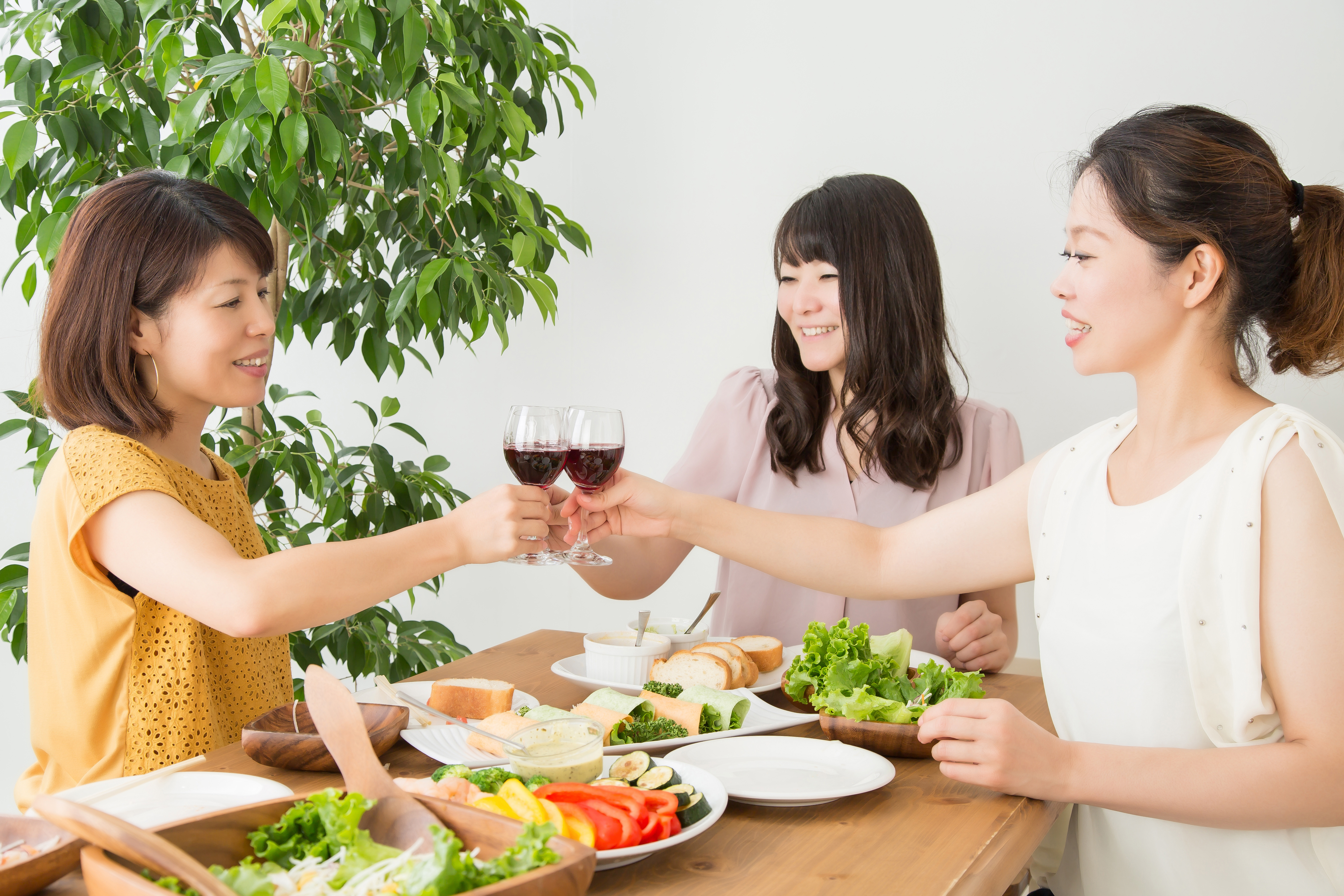 A trio of women from Asian descent cheers with red wine glasses paired with a spread of salads, sandwiches, and fresh vegetables