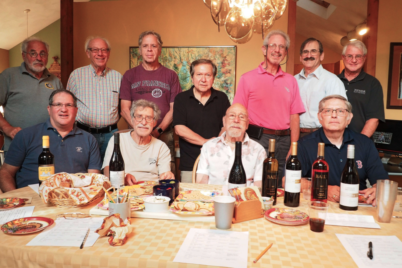 Older men seated and standing around a table loaded with various wine bottles