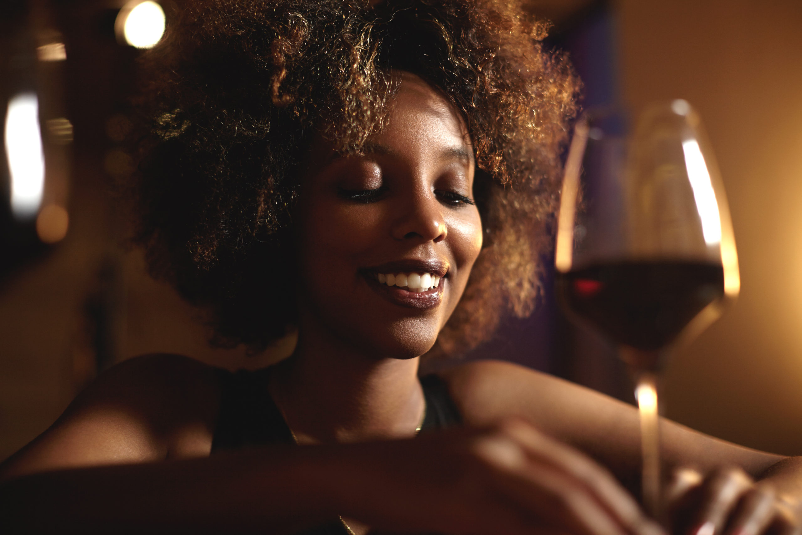Headshot of good-looking young dark-skinned woman with Afro haircut, proposing toast to her friend on birthday party, looking down with shy smile, trying to find right words, holding glass of red wine